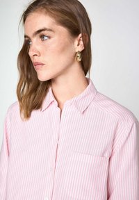 Chemise à rayures rose clair avec un col, une poche avant et une coupe décontractée. Le mannequin porte une boucle d'oreille en or.