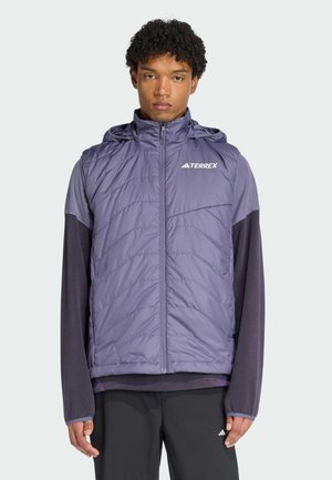 Ung mand iført en lilla quiltet vest med hætte over en langærmet skjorte og sorte bukser, stående mod en ensartet lys baggrund.