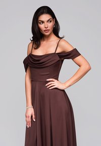Kvinna med mörkt hår i en brungul off-shoulder klänning, guldörhängen och armband, poserar med ena handen på höften mot en enkel bakgrund.