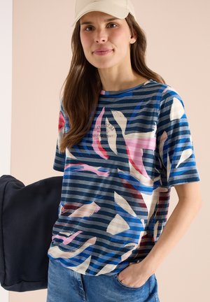 Femme portant un t-shirt à rayures abstraites multicolores, une casquette beige et un jean bleu, tenant une veste sombre dans une main, sur un fond neutre.