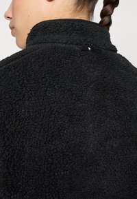 Zwarte fleecejack met een gestructureerd, donzig oppervlak en een hoge kraag. Heeft een klein label bij de nek voor branding.