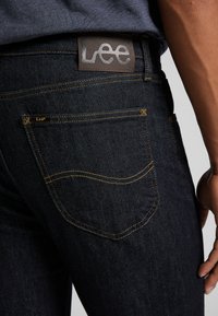 Jeans de mezclilla azul oscuro con costuras amarillas, que presentan un parche de cuero marrón con la etiqueta "Lee" en la cintura y un detalle en el bolsillo trasero.