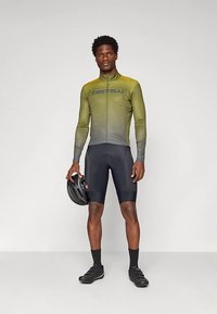 Sportoló zöld árnyalatú hosszú ujjú kerékpáros mezben, fekete párnázott rövidnadrágban, fekete zokniban és cipőben, kezében fekete kerékpáros sisakkal.