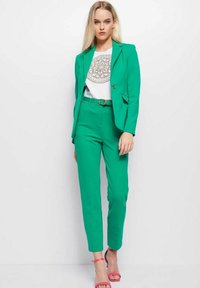Groen op maat gemaakt pak met slim-fit broek en blazer, gecombineerd met een witte graphic t-shirt en roze strappy heels. Gladde textuur, op maat gemaakte ontwerp.