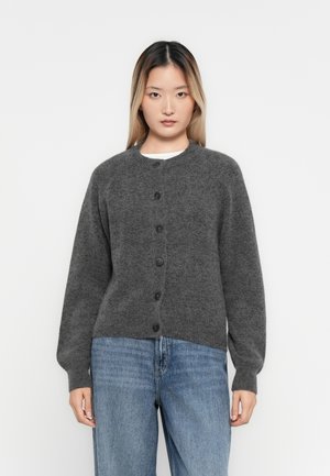 Vrouw in een donkergrijze knopen cardigan met lange mouwen en een blauwe spijkerbroek, staand tegen een effen witte achtergrond.