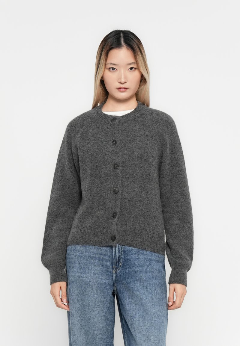 Frau mit einer dunkelgrauen knöpfbaren Strickjacke mit langen Ärmeln und blauen Jeans, die vor einem schlichten weißen Hintergrund steht.
