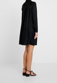 Robe chemise noire plissée avec des manches longues et un col pointu. Fabriquée en tissu lisse, elle présente des poignets boutonnés et une coupe décontractée.