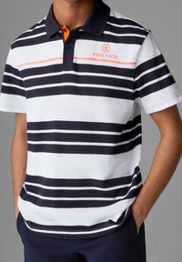 Chemise polo à rayures horizontales bleu marine et blanches avec un accent orange au col. Le matériau semble lisse ; logo sur la poitrine gauche.