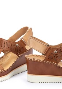 Sandales à talon compensé en cuir brun avec un motif perforé, bout ouvert, sangle ajustable et semelle en caoutchouc blanche. Présente des détails de couture visibles.