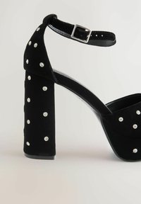 Juodos aukštakulnės sandalai su kulkšnies dirželiu ir sidabriniais kalnų krištolo papuošalais ant kulno ir priekinių dirželių prieš šviesų foną.