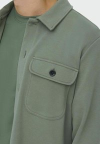 Chaqueta camisa verde claro con un diseño texturizado y acanalado, que incluye un bolsillo en el pecho con botón negro y cuello con solapas.