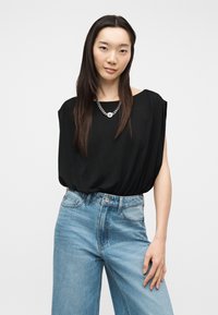 Γυναίκα με μακριά σκουρόχρωμα μαλλιά φορά χαλαρό μαύρο αμάνικο τοπ, ασημένιο αλυσίδα κολιέ και ανοιχτό μπλε τζιν ψηλόμεσο, στέκεται μπροστά σε λευκό φόντο.