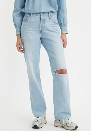 Personne portant un jean droit bleu clair avec une déchirure au genou, une chemise à manches longues bleu clair et des baskets argentées blanches.