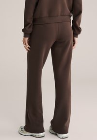 Személy barna egyenes szárú nadrágot és hozzáillő pulóvert visel, hátával a kamera felé áll, fehér tornacipő látható.