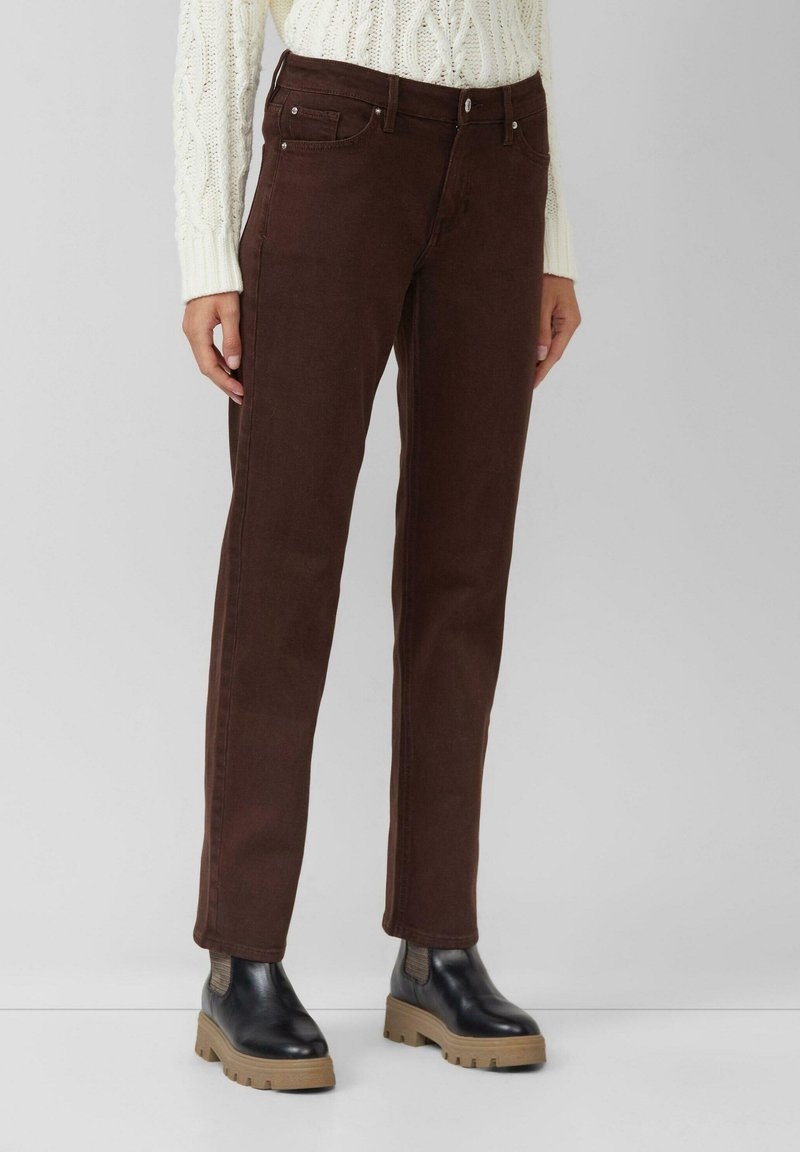 Jean en denim marron avec une coupe droite, cinq poches, fermeture à bouton et couture contrastante, porté avec des bottines noires.