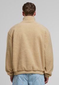 Veste en polaire beige avec un col haut, des poignets et un ourlet côtelés. La texture est douce et duveteuse, offrant une apparence chaleureuse.
