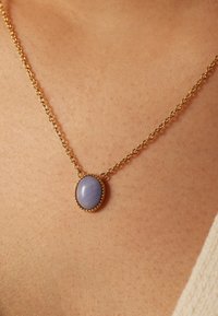 Gouden ketting met een ovale lichtpaarse stenen hanger, omgeven door een gouden rand van kralen, rustend op een gestructureerd huidoppervlak.