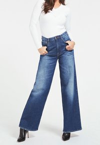 Jeans in denim a gamba larga con lavaggio blu scuro, alta vita, con tasche frontali. Abbinate a un top aderente a maniche lunghe bianco e stivali neri con tacco.