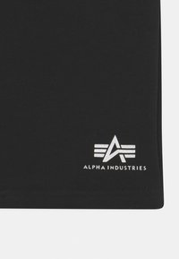 Crna pamučna majica s bijelim logotipom Alpha Industries koji ima tri horizontalne pruge i oštar dizajn na rubu.
