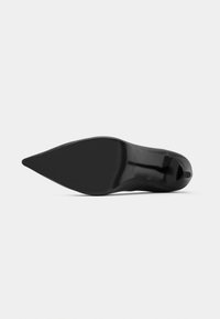 Μαύρο ψηλοτάκουνο παπούτσι με μυτερή μύτη και λείο επάνω μέρος. Η σόλα είναι επίπεδη και υφασμένη, με ελάχιστα διακριτικό λογότυπο.