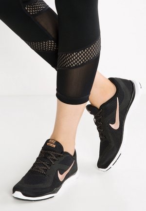 Beine, die schwarze Leggings mit Mesh-Einsätzen und schwarze Nike Laufschuhe mit pinkem Swoosh und weißen Sohlen auf einer weißen Oberfläche tragen.