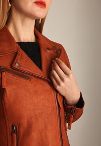 Kvinde justerer kraven på en rustfarvet læderjakke, iført en sort tekstureret top og guldøreringe, på en neutral baggrund.