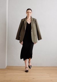 Oliven grønn oversized blazer over en svart ärmeløs kjole med buet nederkant og spisse svarte sko, satt mot en nøytral bakgrunn.
