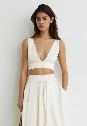 Pikkade tumedate juustega naine, kes kannab valget varrukateta sügava V-kaelusega lühikest topi ja kõrge vöökohaga valget volditud seelikut külje paelaga, seisab valge seina taustal.