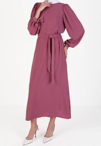 Robe longue mauve avec manches bouffantes, ceinture assortie et tissu texturé. Comprend un col rond et une silhouette fluide, associée à des talons blancs.
