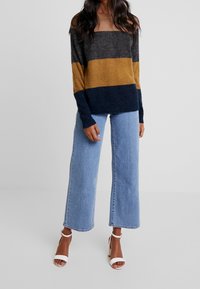 Color-blocked sweater in dark gray, mustard, and navy; soft texture, wide neckline, paired with light blue wide-leg jeans and white heels.