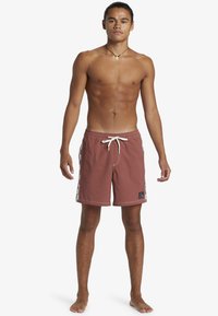 Red swim trunks made of soft fabric, featuring a white drawstring, checkerboard side pattern, and a logo patch on the hem.