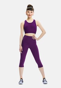 Young woman in purple athletic crop top and capri leggings stands confidently with hand on hip and black sneakers on white background.
