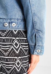 Giacca di jeans azzurro chiaro presentata con bottoni a pressione argentati; abbinata a una gonna con motivo geometrico in bianco e nero caratterizzata da forme audaci.