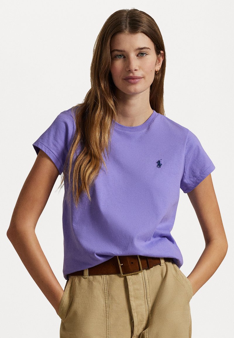T-shirt di cotone viola con collo a giro, maniche corte e un piccolo logo blu navy sul petto. Abbinato a pantaloni kaki chiari e una cintura marrone.