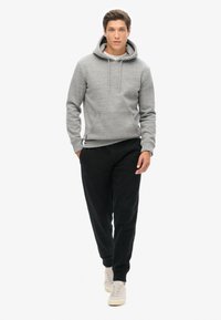 Grijze hoodie met trekkoorden, kangoeroe-pocket, gedragen met zwarte joggers en lichtgekleurde sneakers. Glad textuur, ontspannen pasvorm.