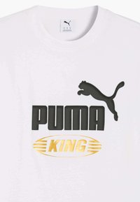 Maglietta bianca a girocollo con logo Puma nero e testo sopra l'emblema dorato "KING" incorniciato da linee ovali stilizzate al centro davanti.