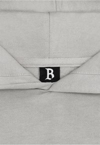Grau Hoodie aus strukturiertem Stoff, mit einem schwarzen Etikett mit einem weißen Buchstaben "B" am Hals. Einfaches, minimalistisches Design.