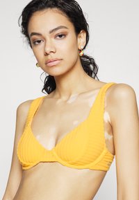 Gul ribbet bikini topp med brede stropper og trekantet koppdesign. Har teksturert stoff og en liten logodetalj på siden.