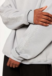 Světlé šedé mikina s žebrovanými manžetami, která má vyražený bílý nápis na rukávech a logo na spodním lemu. Měkká textilie.