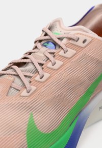 Tangy beige szövetű sneaker közeli képe, rózsaszín fűzőkkel, zöld és kék Nike emblémával az oldalon és a nyelvén.