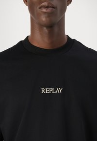 Černé bavlněné tričko s kulatým výstřihem a kontrastním vyšívaným logem "REPLAY" v béžové barvě na levém prsu.