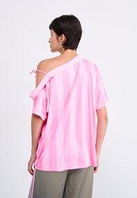 Rosa randig oversize skjorta med korta ärmar, med en off-shoulder-design och en knutdetalj på ena axeln. Mjuk, slät textur.