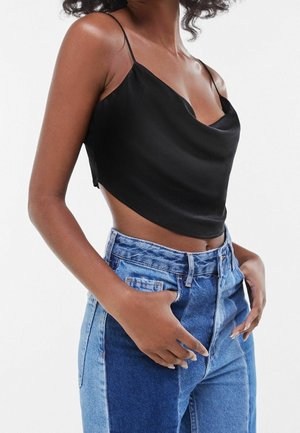 Frau trägt ein schwarzes, satiniertes, bauchfreies Camisole und hoch taillierte, zweifarbige blaue Jeans mit Händen in den Taschen.