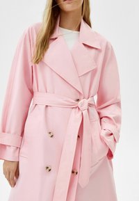 Light pink trench coat with a belted waist, large lapels, and double-breasted button closure. Features long sleeves and side pockets.