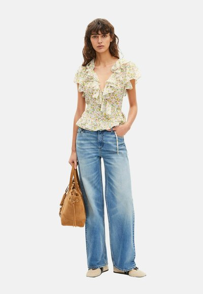 Mujer con blusa floral con volantes y mangas cortas, vaqueros azules de pierna ancha, zapatos beige y sujetando un bolso de ante marrón.