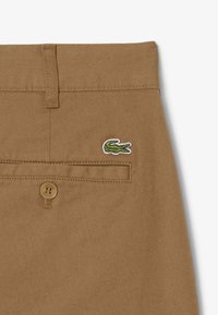 Pantalons kaki avec une poche arrière à bouton, un tissu texturé subtil et un logo crocodile brodé en vert et blanc.