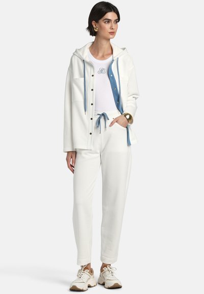 Femeie purtând hanorac alb cu șnururi albastre, pantaloni albi cu șnur albastru, adidași albi și brățară aurie, stând cu mâna în buzunar.