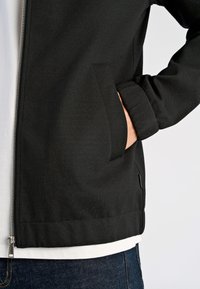 Czarna kurtka na zamek z gładką fakturą, bocznymi kieszeniami i luźnym krojem, noszona na białej koszuli i niebieskim denimie.