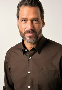 Ein mittelalter Mann mit kurzen dunklen Haaren und Bart trägt ein braunes Button-up-Hemd und blickt direkt in die Kamera vor einem schlichten Hintergrund.