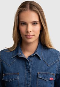 Camisa de denim en azul con costuras de color naranja claro, botones a presión, detalle de yugo puntiagudo y dos bolsillos en el pecho con etiqueta de logo a la derecha.